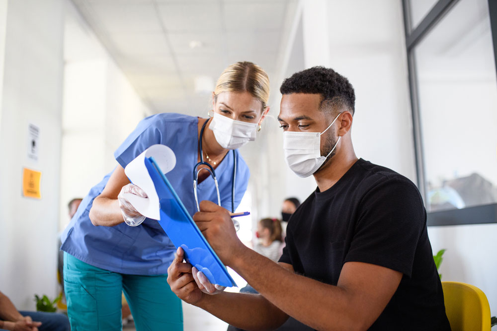 man wearing mask with clipboard talking to doctor